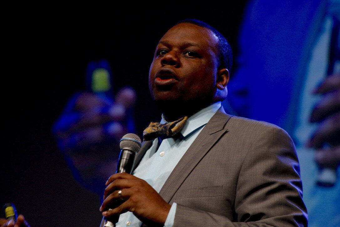 Adam speaking into a microphone on stage in a gray suit and gold bow tie